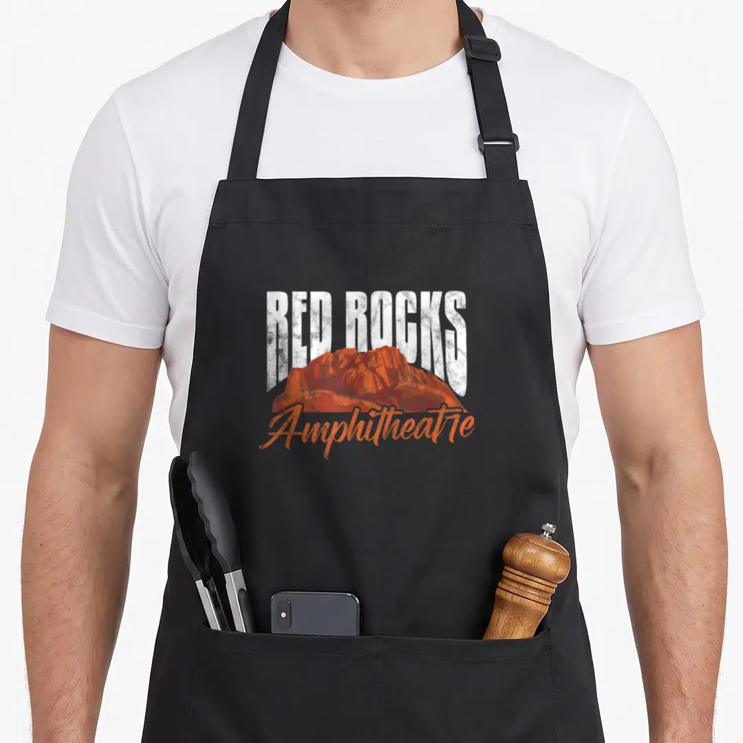 Amphitheatre morrisons apron - Distressed white RED ROCKS text over orange rock formation with orange Amphitheatre — shown on a black cotton canvas apron against a white background