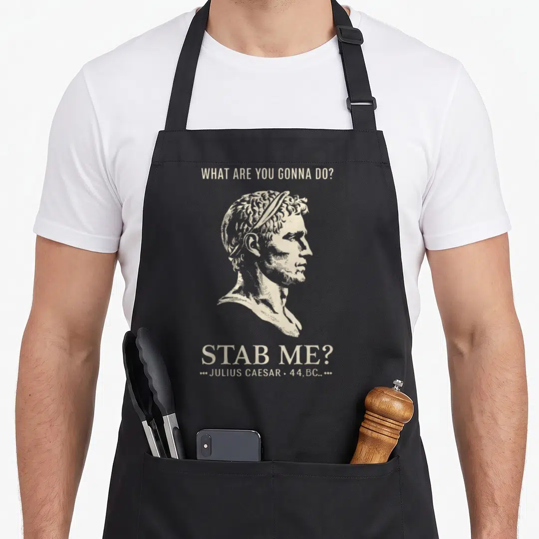 Julius meme apron - Beige profile bust of Julius Caesar with sarcastic stab me quote text — shown on a black cotton canvas apron against a white background