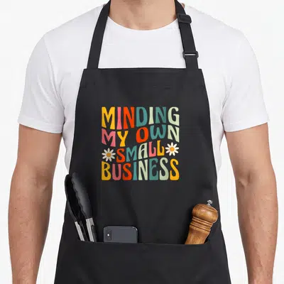 Minding owner entrepreneur small business aprons - Stacked retro text MINDING MY OWN SMALL BUSINESS with two flowers — shown on a black cotton canvas apron against a white background