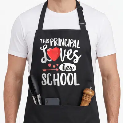Principal valentine's day apron - White text This Principal Loves her School with red heart replacing O and arrow above — shown on a black cotton canvas apron against a white background