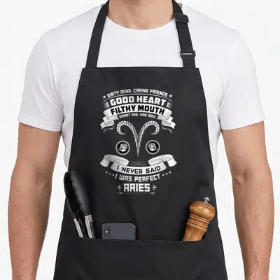 Ary zodiac birthday apron - White ram horns and stacked ribbons with sarcastic text on black fabric — shown on a black cotton canvas apron against a white background
