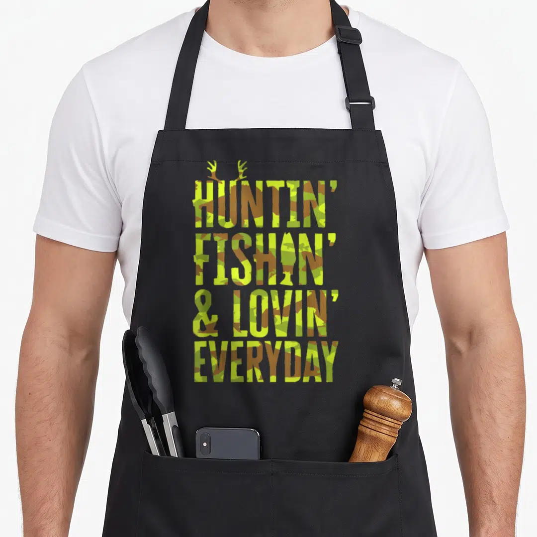 Hunting camo apron - Stacked camo text reading Huntin Fishin Lovin Everyday with antlers above — shown on a black cotton canvas apron against a white background