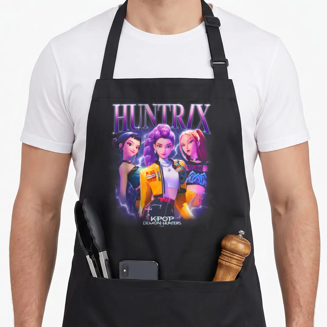Huntr x apron - Three women with HUNTR/X text and blue lightning in purple yellow blue pink and white — shown on a black cotton canvas apron against a white background