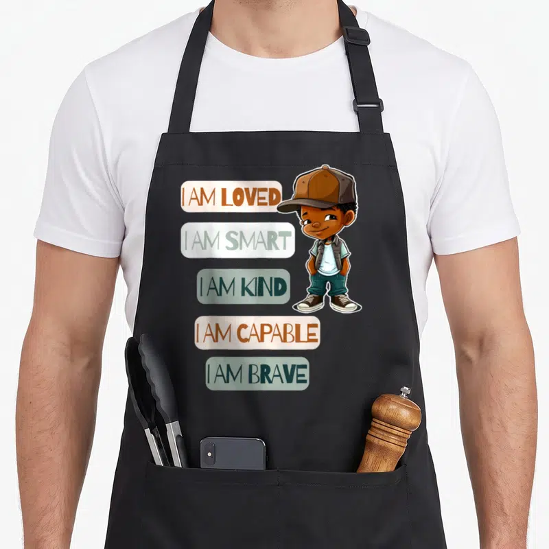 Positive african american aprons - Cartoon boy beside five banners reading I AM LOVED SMART KIND CAPABLE BRAVE in brown — shown on a black cotton canvas apron against a white background