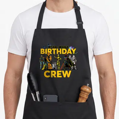 Character crew birthday apron - STAR WARS BIRTHDAY CREW text with cartoon characters and spacecraft in yellow black — shown on a black cotton canvas apron against a white background