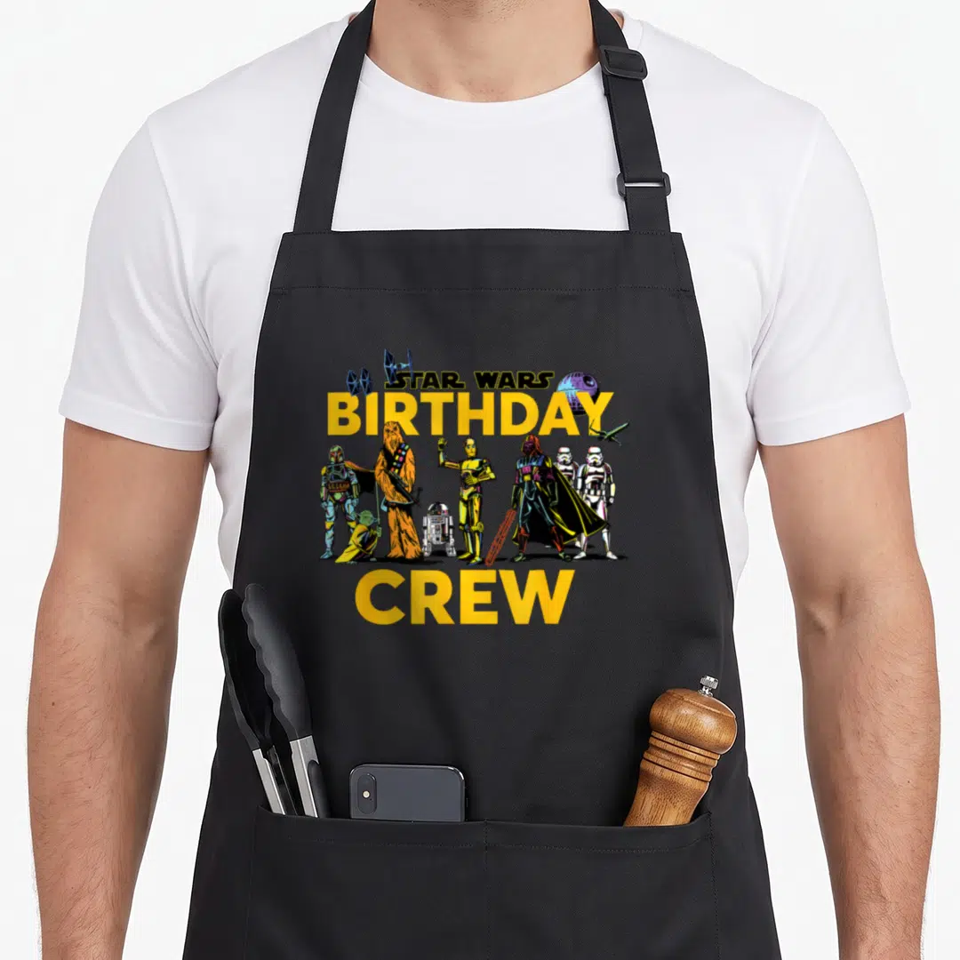 Character crew birthday apron - STAR WARS BIRTHDAY CREW text with cartoon characters and spacecraft in yellow black — shown on a black cotton canvas apron against a white background