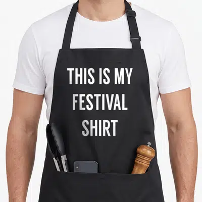 Music festival apron - White stacked text reading THIS IS MY FESTIVAL SHIRT on grey background — shown on a black cotton canvas apron against a white background