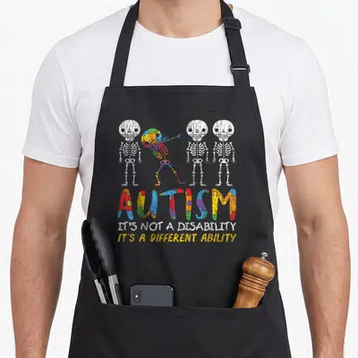 Autism skeleton apron - Four skeletons including one multi-colored dabber above text reading Autism It's Not — shown on a black cotton canvas apron against a white background