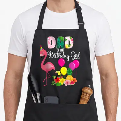 Funny birthday flamingo apron - Dad of the Birthday Girl text with cartoon flamingo, toucan, balloons, and tropical — shown on a black cotton canvas apron against a white background