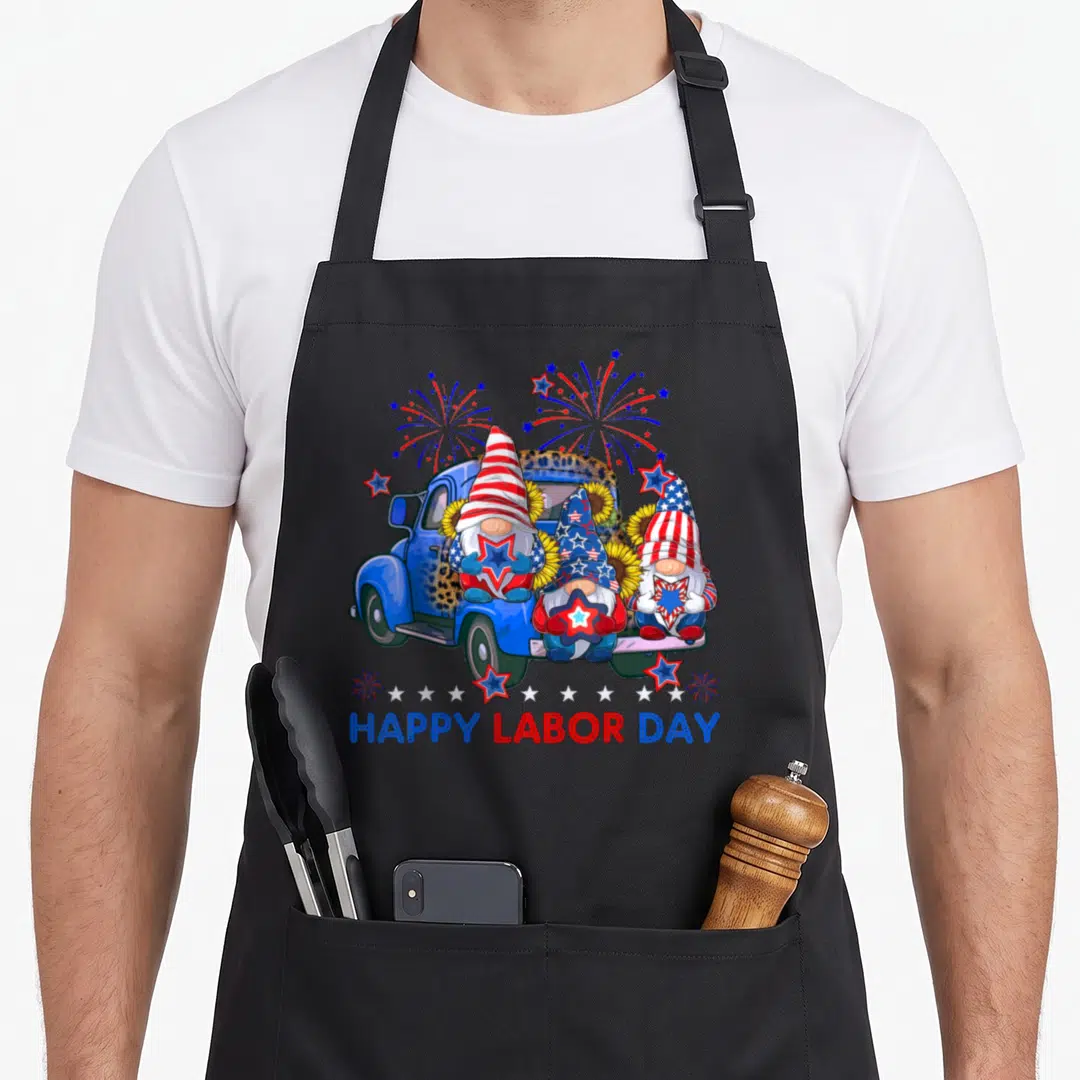 Firework american flag apron - Three gnomes in blue truck holding stars with fireworks and HAPPY LABOR DAY text — shown on a black cotton canvas apron against a white background