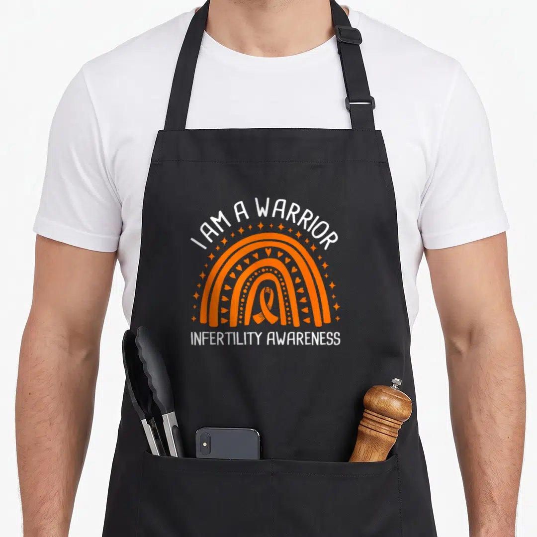 Funny warrior infertility awareness apron - Orange rainbow with ribbon and hearts under I AM A WARRIOR text — shown on a black cotton canvas apron against a white background