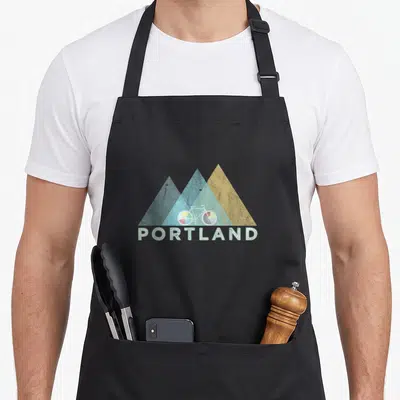 Portland bicycle apron - Three blue and yellow mountain triangles with white bike and PORTLAND text — shown on a black cotton canvas apron against a white background