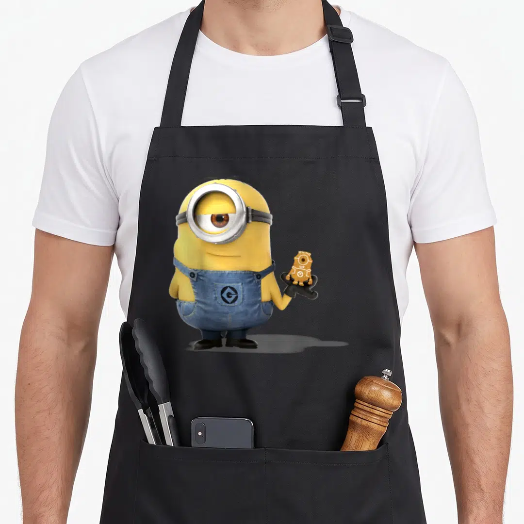 Despicable bread apron - Yellow Minion in blue overalls holding a gingerbread cookie portrait — shown on a black cotton canvas apron against a white background