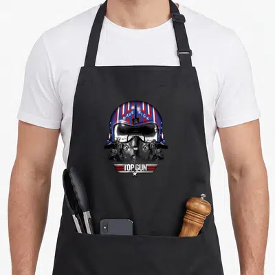 Helmet gun apron - Realistic flight helmet with MAVERICK text and TOP GUN winged banner in blue red white black — shown on a black cotton canvas apron against a white background