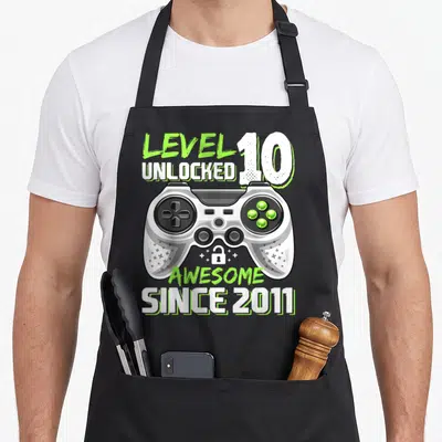 10th birthday video game apron - LEVEL 10 UNLOCKED game controller with AWESOME SINCE 2011 text and lock icon — shown on a black cotton canvas apron against a white background