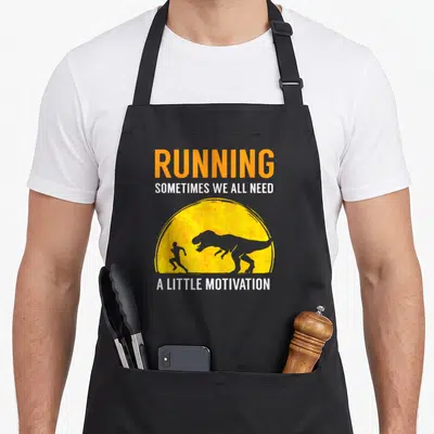 Running jogging apron - T-Rex chasing runner in yellow sun with Running Sometimes We All Need A Little Motivation text — shown on a black cotton canvas apron against a white background