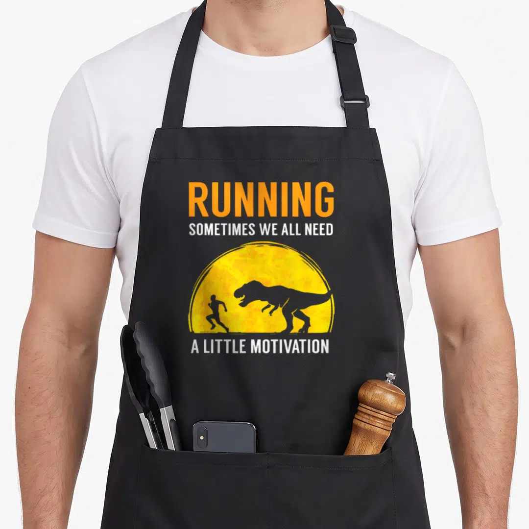 Running jogging apron - T-Rex chasing runner in yellow sun with Running Sometimes We All Need A Little Motivation text — shown on a black cotton canvas apron against a white background