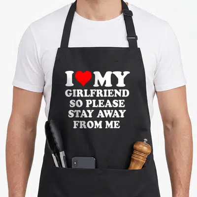 Stay girlfriend funny apron - White text I MY GIRLFRIEND SO PLEASE STAY AWAY FROM ME with a red heart replacing — shown on a black cotton canvas apron against a white background