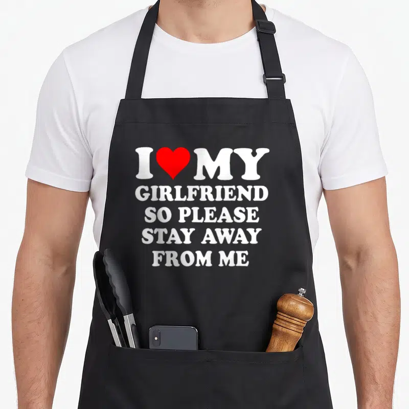 Stay girlfriend funny apron - White text I MY GIRLFRIEND SO PLEASE STAY AWAY FROM ME with a red heart replacing — shown on a black cotton canvas apron against a white background