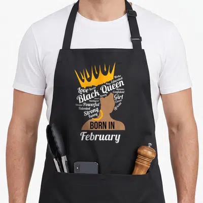 Strong queen birthday apron - Crowned woman silhouette with hair made of positive words and Born In February text — shown on a black cotton canvas apron against a white background