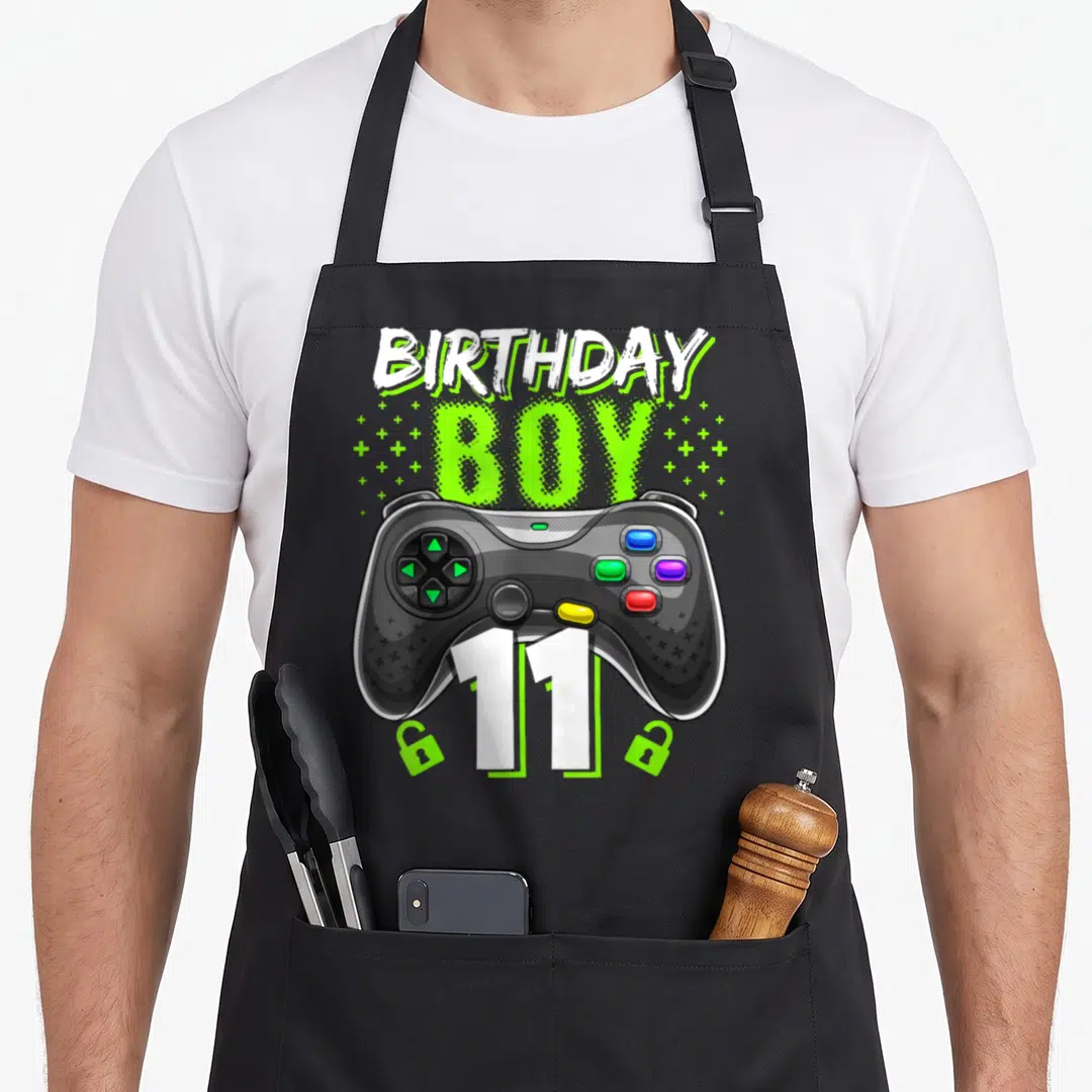 Controller gamer video game apron - Cartoon controller with BIRTHDAY BOY 11 text surrounded by green plus signs and dots — shown on a black cotton canvas apron against a white background