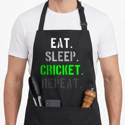 Funny cricket apron - Stacked white grey and green stencil text reading Eat Sleep Cricket Repeat — shown on a black cotton canvas apron against a white background