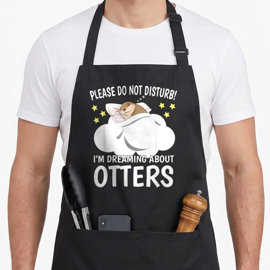 Dreaming otter mom apron - Sleeping brown otter on pink pillow under white blanket with yellow stars and white text — shown on a black cotton canvas apron against a white background
