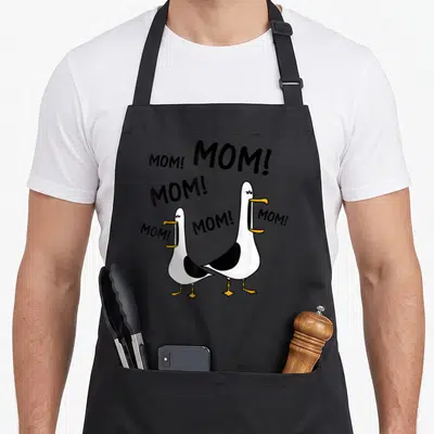 Seagull mama mother's day aprons - Two cartoon seagulls with open beaks surrounded by scattered MOM! text in black — shown on a black cotton canvas apron against a white background