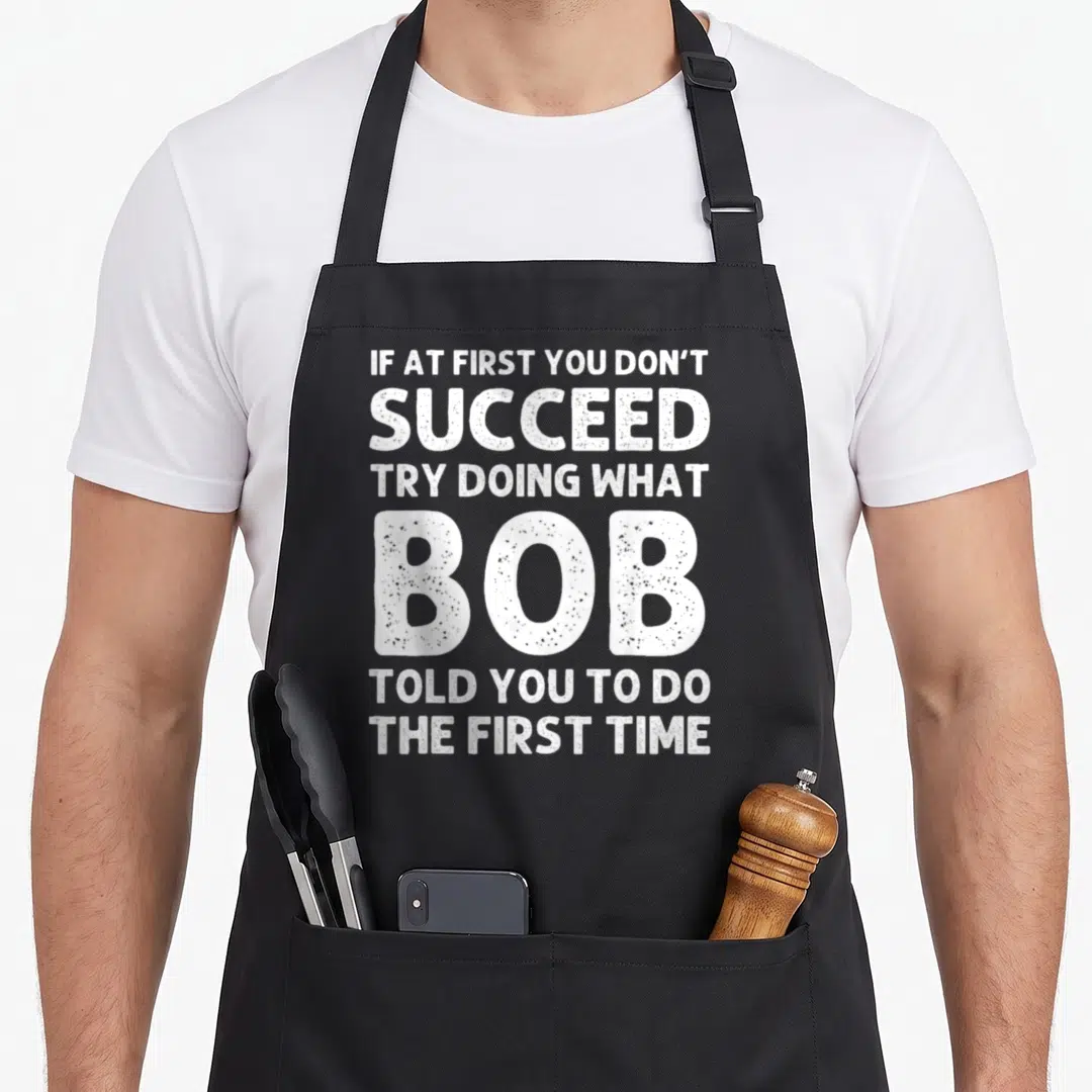 Bob christmas apron - White distressed text IF AT FIRST YOU DON'T SUCCEED TRY DOING WHAT BOB TOLD YOU TO DO THE FIRST — shown on a black cotton canvas apron against a white background