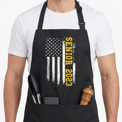 Senior graduation american flag apron - Distressed white flag with vertical yellow SENIOR 2023 text on black apron — shown on a black cotton canvas apron against a white background