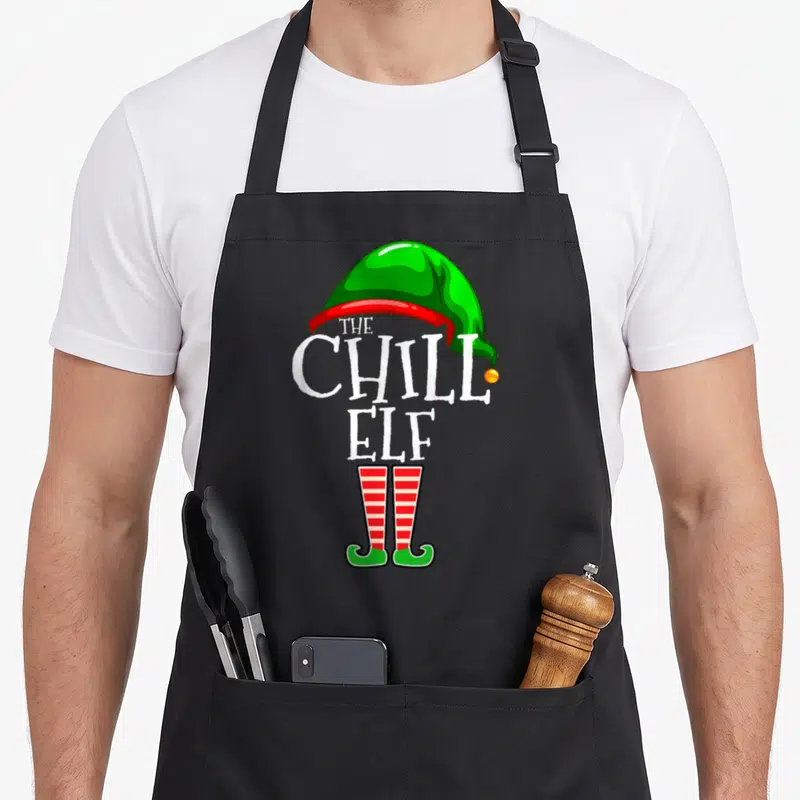 Chill family christmas elf apron - Green elf hat, white text reading The Chill Elf, and red striped shoes in cartoon — shown on a black cotton canvas apron against a white background