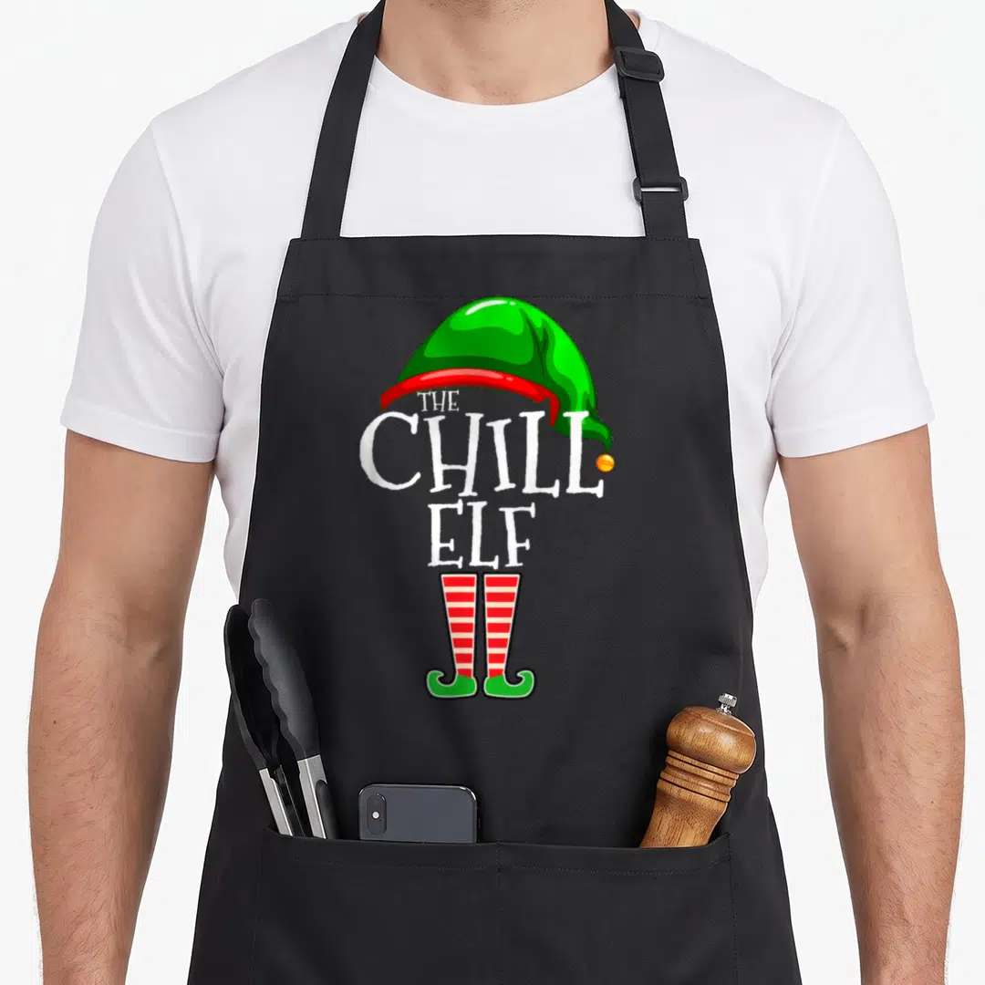 Chill family christmas elf apron - Green elf hat, white text reading The Chill Elf, and red striped shoes in cartoon — shown on a black cotton canvas apron against a white background