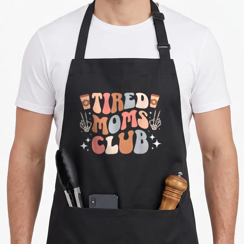 Exhausted mother's apron - Retro text reading TIRED MOMS CLUB with coffee cups and skeleton hands in pastel colors — shown on a black cotton canvas apron against a white background