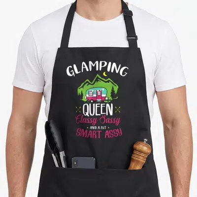 Glamping smart apron - Camper van scene with Glamping Queen Classy Sassy text in white pink and green — shown on a black cotton canvas apron against a white background