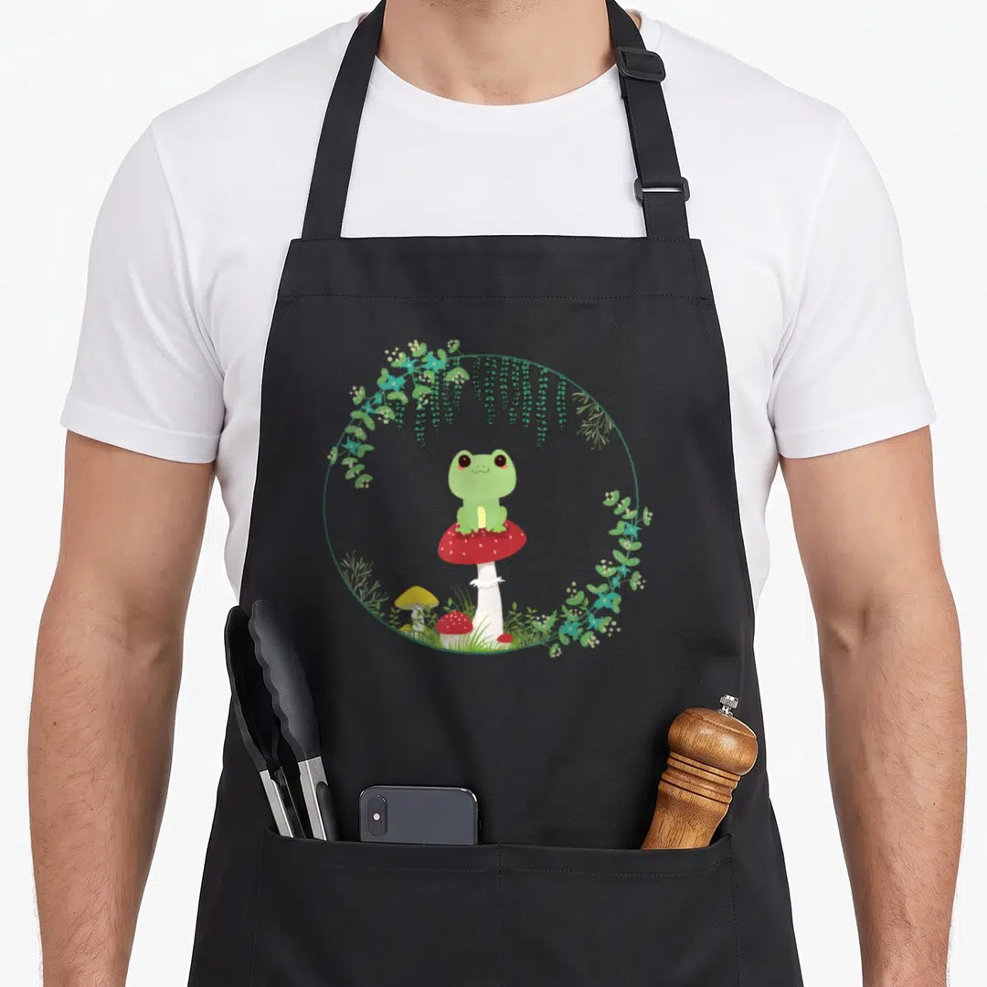 Kawaii frog cottagecore apron - Green cartoon frog sits on red mushroom with white dots inside circular wreath — shown on a black cotton canvas apron against a white background
