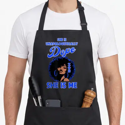 Zeta pride apron - Cartoon woman with afro, ZΦB in hair, blue text reading SHE IS UNAPOLOGETICALLY Dope SHE IS ME — shown on a black cotton canvas apron against a white background