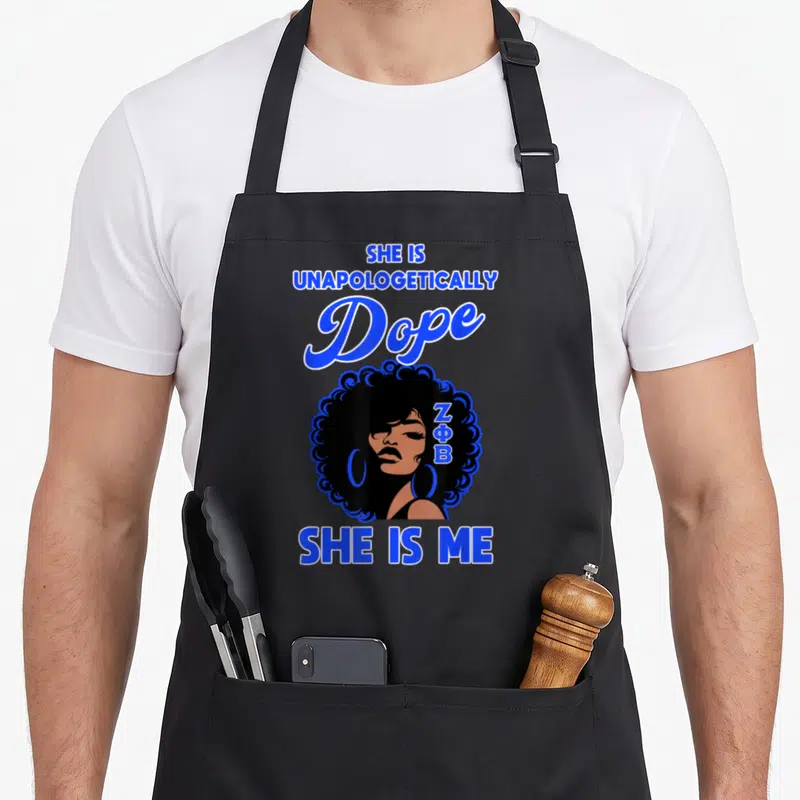 Zeta pride apron - Cartoon woman with afro, ZΦB in hair, blue text reading SHE IS UNAPOLOGETICALLY Dope SHE IS ME — shown on a black cotton canvas apron against a white background
