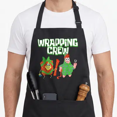 Wrapping christmas apron - SpongeBob wrapped in red paper and Patrick in green paper holding scissors below text — shown on a black cotton canvas apron against a white background