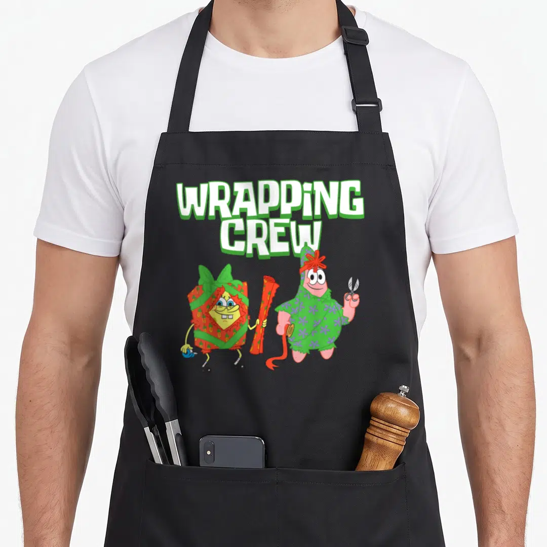 Wrapping christmas apron - SpongeBob wrapped in red paper and Patrick in green paper holding scissors below text — shown on a black cotton canvas apron against a white background