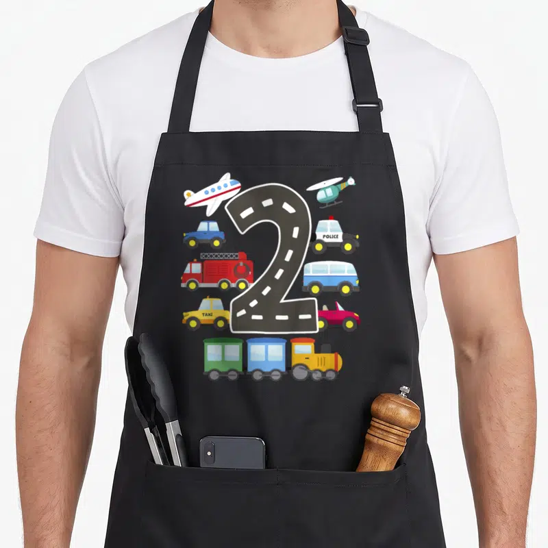 Transportation 2nd apron - Number two road shape surrounded by cartoon police car, taxi, train, plane, and helicopter — shown on a black cotton canvas apron against a white background