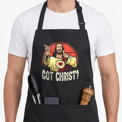 Buddy christmas apron - Cartoon Jesus winking thumbs up red halo GOT CHRIST text yellow robe — shown on a black cotton canvas apron against a white background