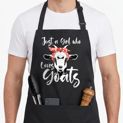 Funny goat aprons - Cartoon goat head with red polka dot bandana and white brush script text — shown on a black cotton canvas apron against a white background
