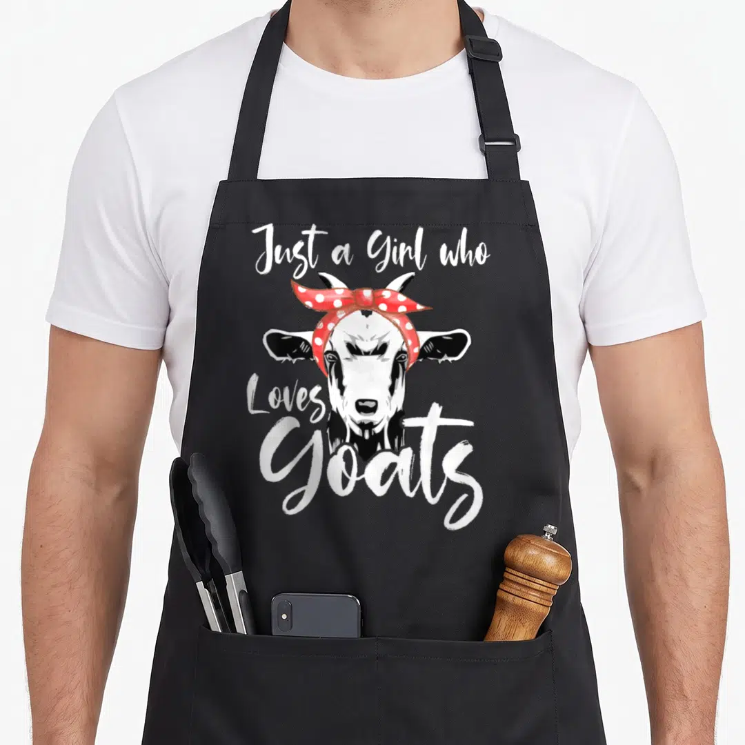 Funny goat aprons - Cartoon goat head with red polka dot bandana and white brush script text — shown on a black cotton canvas apron against a white background