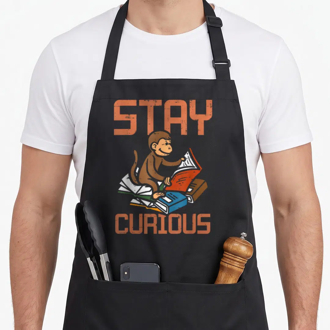 Stay librarian husband apron - Cartoon monkey reading books with orange STAY CURIOUS text and blue red brown volumes — shown on a black cotton canvas apron against a white background