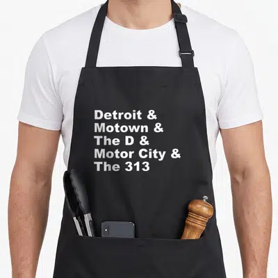 Nickname detroit apron - White stacked text listing Detroit nicknames including Motown Motor City The 313 The D — shown on a black cotton canvas apron against a white background