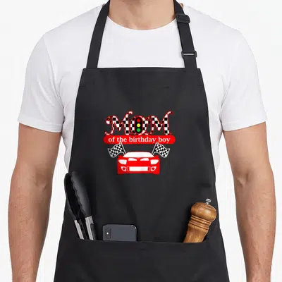 Birthday race car apron - MOM with traffic light O, red banner, red race car, two checkered flags, cartoon style — shown on a black cotton canvas apron against a white background