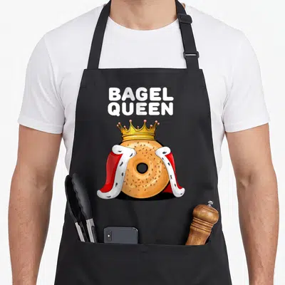 Bagel queen apron - Cartoon tan bagel wearing gold crown and red white cape under white BAGEL QUEEN text — shown on a black cotton canvas apron against a white background