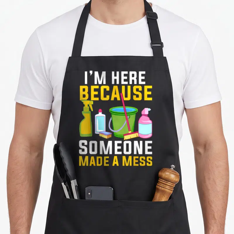 Funny housekeeping housekeeper apron - I'M HERE BECAUSE SOMEONE MADE A MESS text with cartoon bucket broom and bottles — shown on a black cotton canvas apron against a white background