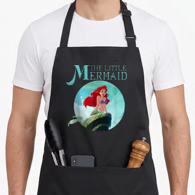 Rock splash aprons - Cartoon mermaid with red hair on rock inside blue circle with stacked text — shown on a black cotton canvas apron against a white background