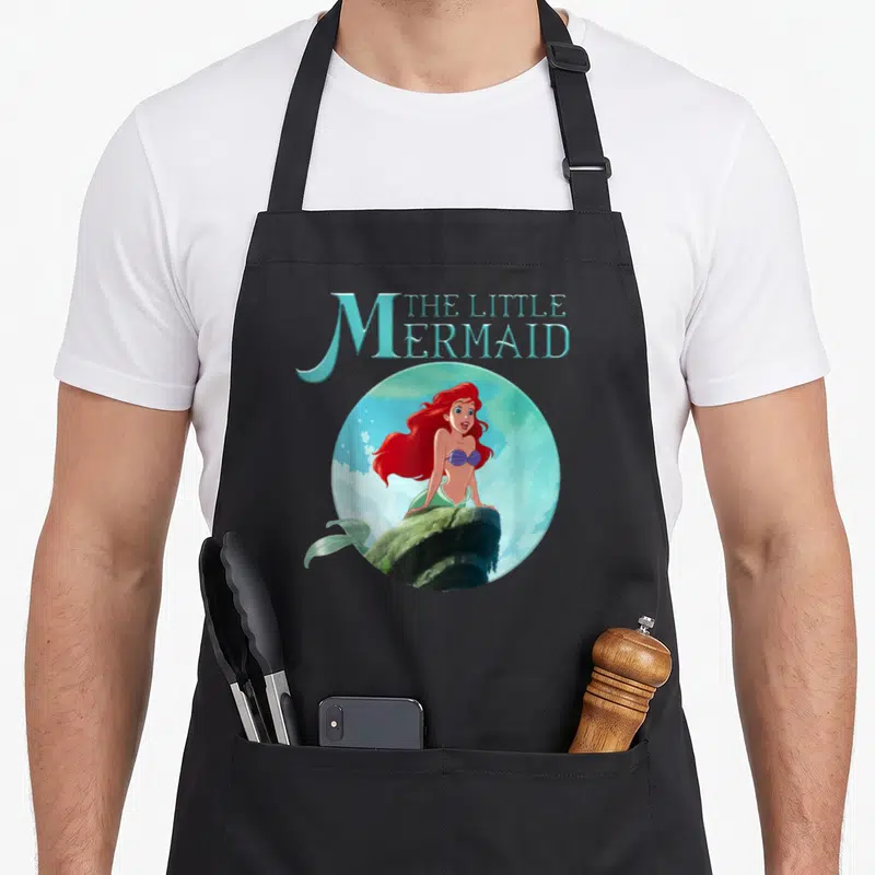 Rock splash aprons - Cartoon mermaid with red hair on rock inside blue circle with stacked text — shown on a black cotton canvas apron against a white background